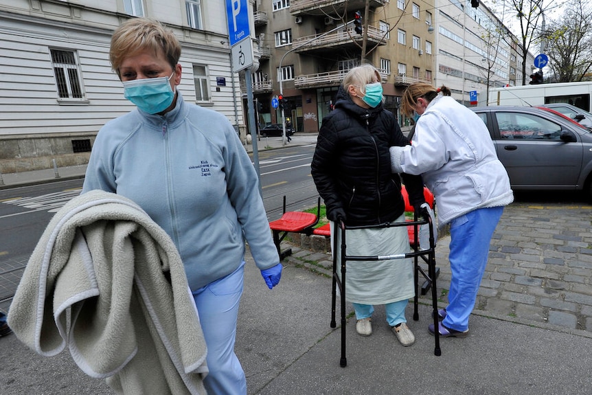 Three women in medical gowns and facemasks walk on a cobblestone path on an overcast day.