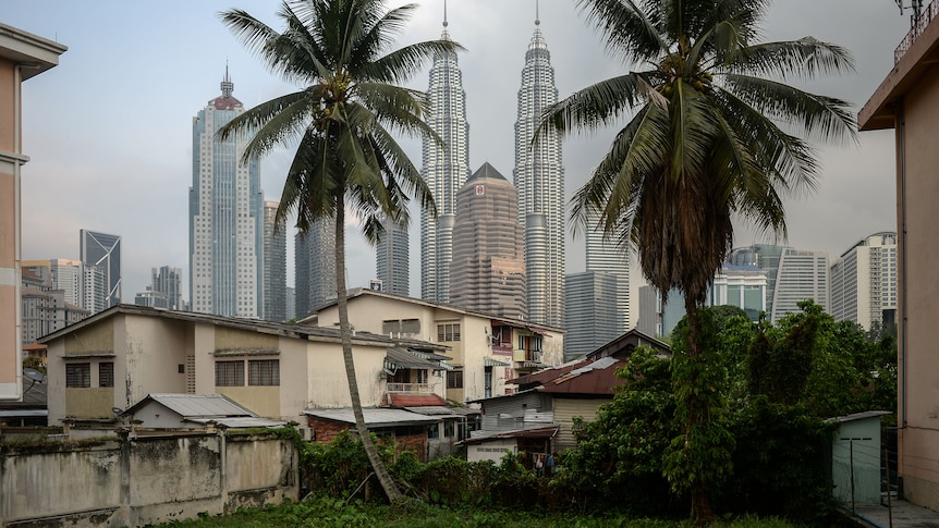 The city skyline of Kuala Lumpur can be seen between a pair of palm trees in a jungle garden.