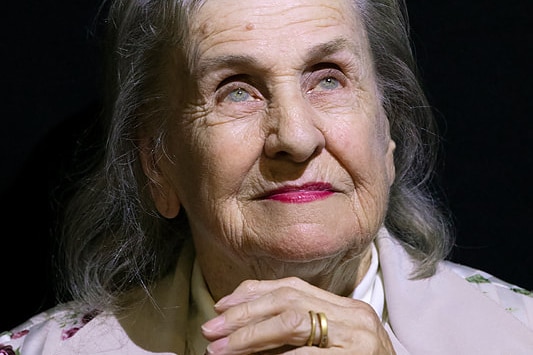 A woman in a pale purprle top with fair eyes and dark grey hair, hands clasped in front of her looking upwards