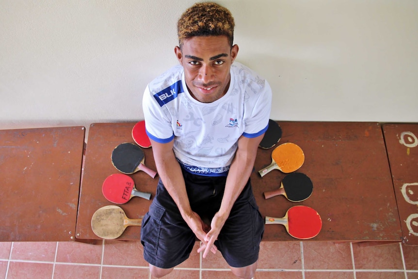 A Fijian man in his early 20s looks up a camera. He sits on a bench surrounded by table tennis paddles.