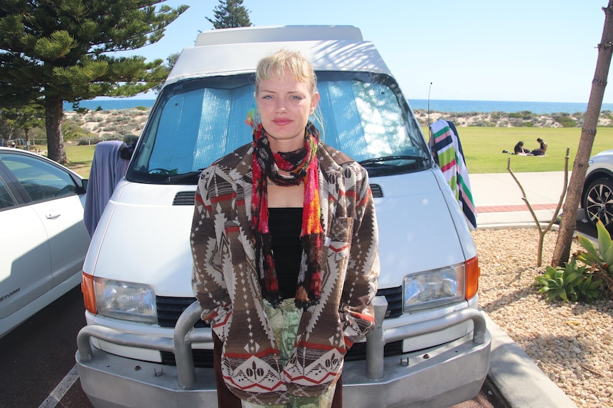 A blonde woman stands in front of a van on a beachfront
