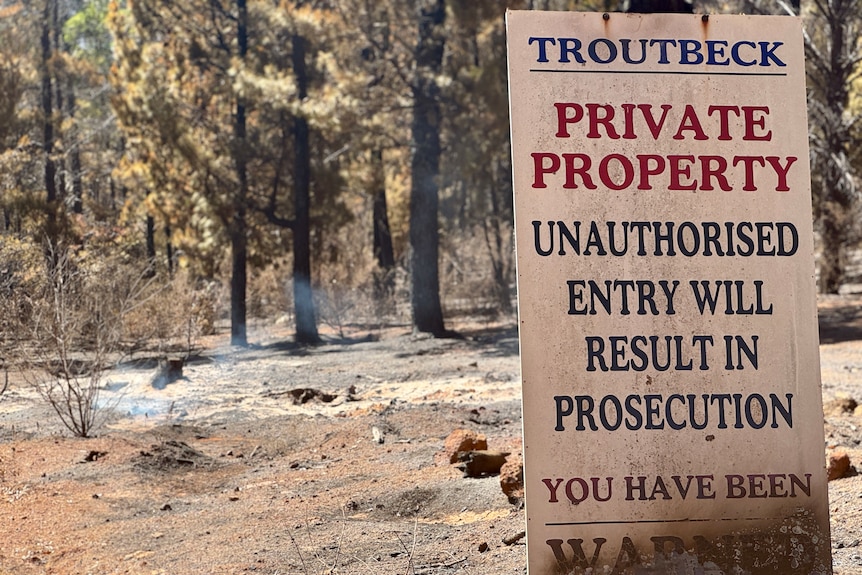 A private property sign stands burnt and charred in the dirt as the background soil and burnt wood remains still in the sun