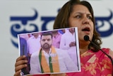 A woman wearing a sari holds up a photo of a man wearing a white shirt and red and yellow scarf.