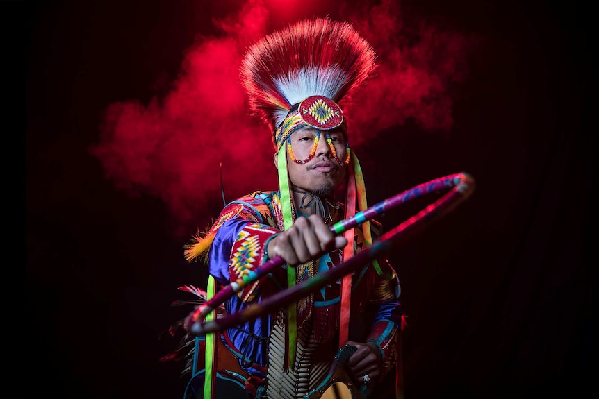 A man in bright coloured Native American regalia and headpiece holds hoop with outstretched arm, stands in front of red smoke.