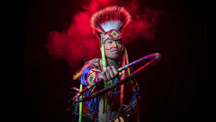 A man in bright coloured Native American regalia and headpiece holds hoop with outstretched arm, stands in front of red smoke.