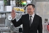 Elon Musk in a black suit stands and waves in front of a metal car part, in a factory.