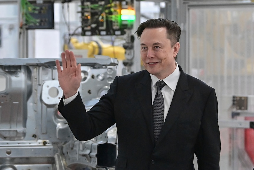 Elon Musk in a black suit stands and waves in front of a metal car part, in a factory.