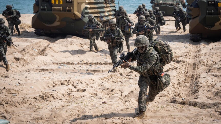 Marines in camouflage gear run onto the beach, between military vehicles