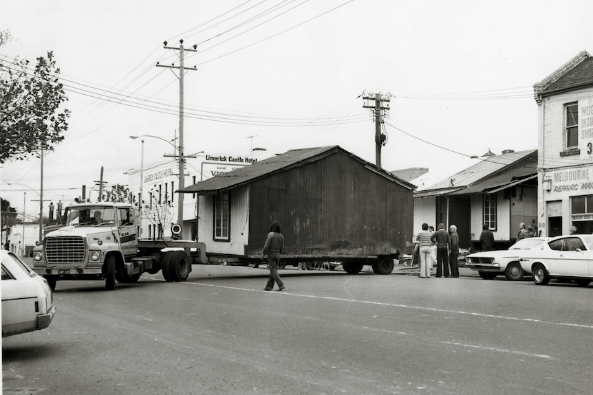 A small iron house is being transported on the back of a truck as bystanders watch on.