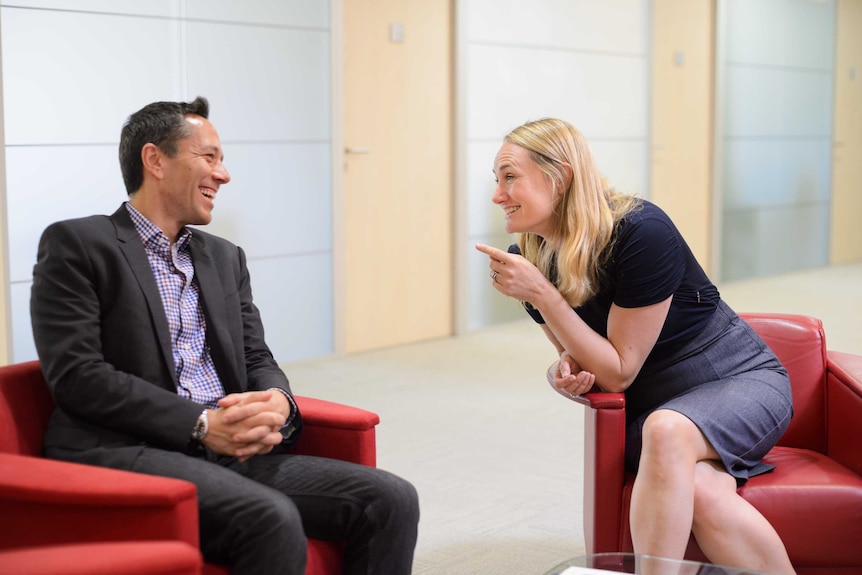 A man and a woman sit in separate red chairs, she points a finger at him as they both laugh.