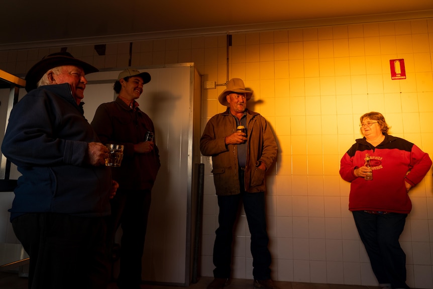 A group of people stand around drinking and laughing in an industrial kitchen while the sun sets.