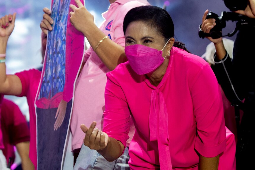 A woman wearing a bright pink mask does a love hand symbol