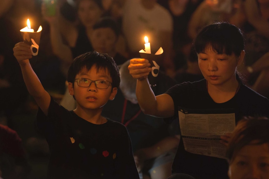 A young boy holds up a candle at night at the vigil next to a woman doing the same.