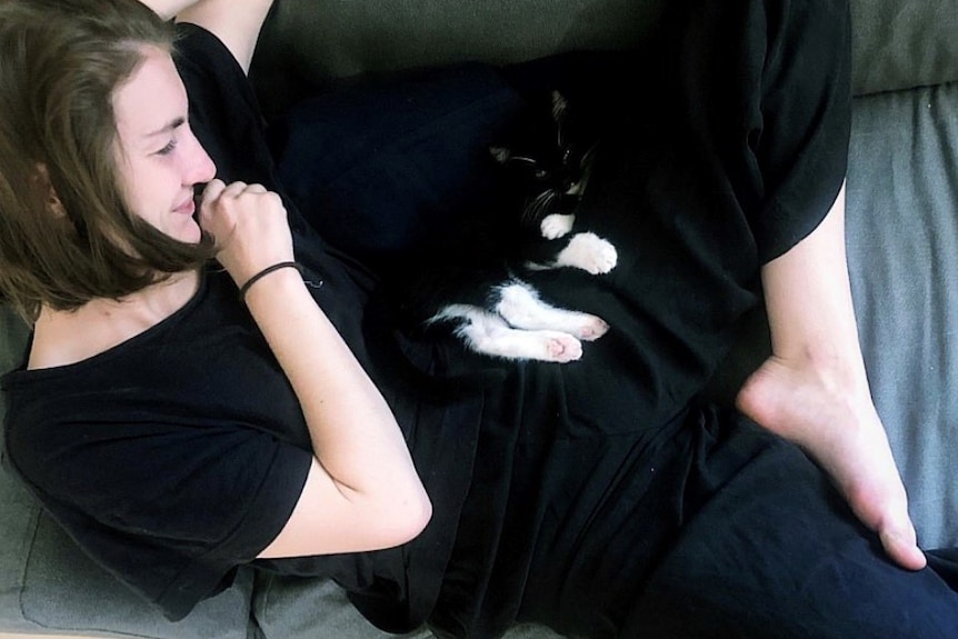 A photo from above showes a woman lying on the couch with a black and white cat in her lap