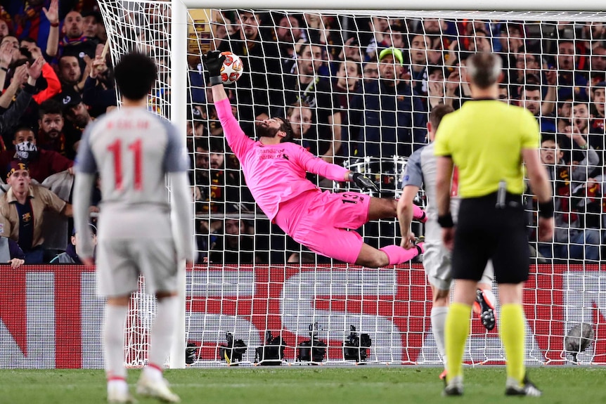 A goalkeeper flies through the air, his hand grasps at a ball that goes past him into the net.