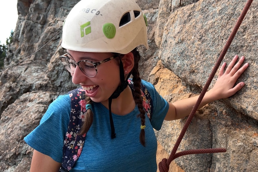 a girl with glasses, a white helmet, blue top and braids smiles and looks down as she prepares to climb.