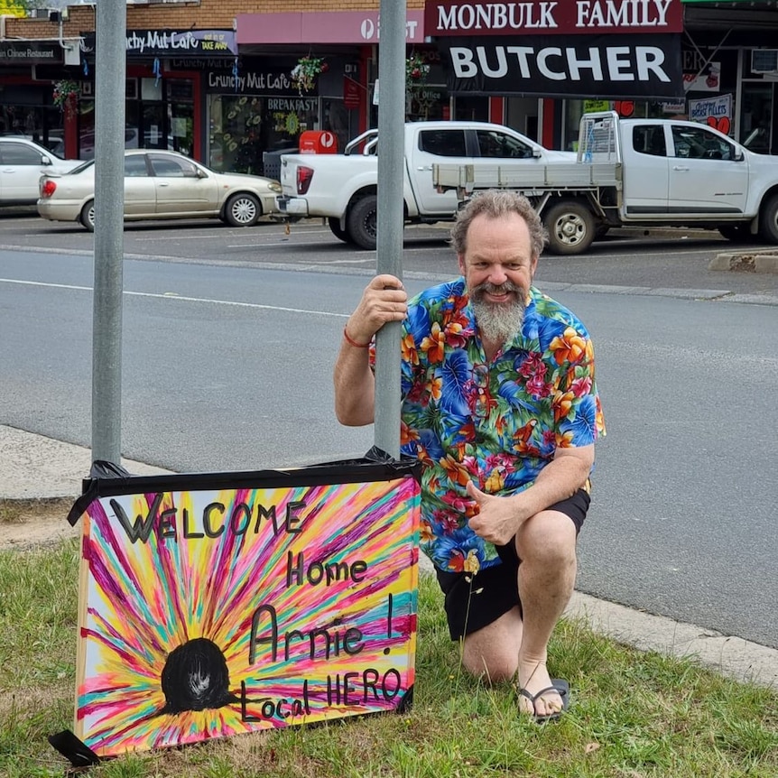 Man kneels next to a sign by a light pole that reads 'Welcome Home Arnold local hero' in colourful writing. Shops in background