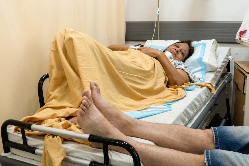 A woman lies on a hospital bed with a yellow sheet draped over her as a man rests his feed on the bed.