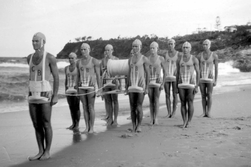 Nine men in 1950s lifesaver uniforms stand in a line on a beach with belt and reel.