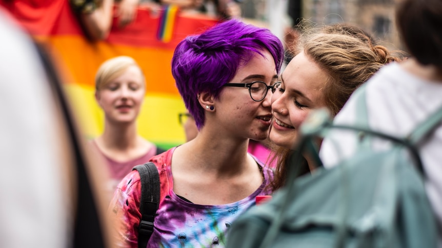 One young girl with short purple hair and glasses kissing the cheek of another adorable girl with a nose ring.
