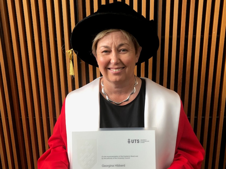 A middle-aged woman stands in a university graduate's gown and cap