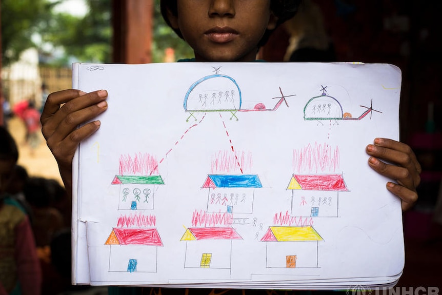 A child holds up a drawing that shows helicopters shooting people's houses.