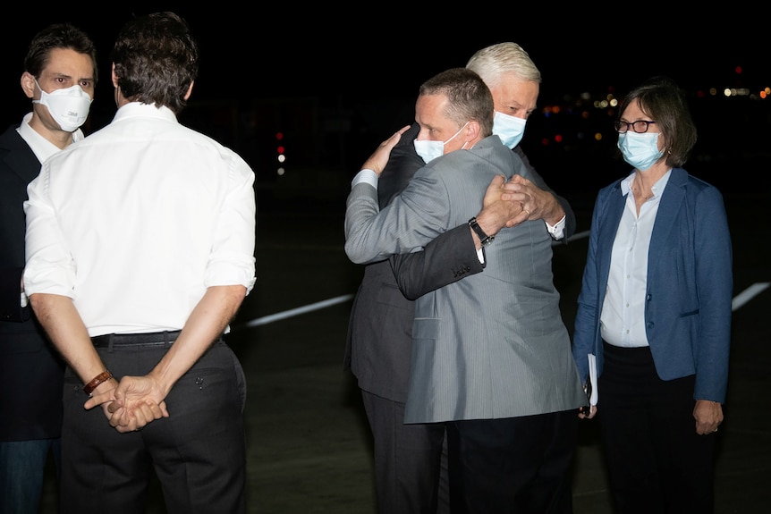 Two men hugging while another two men talk and a woman stands nearby on an airport tarmac.