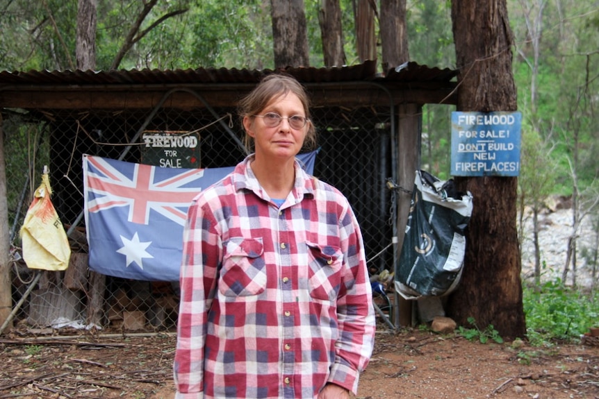 A woman wearing a chequered shirt stands in front of an Australian flag and firewood sign.