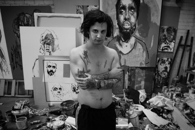 A shirtless artist stands amongst his half-finished paintings