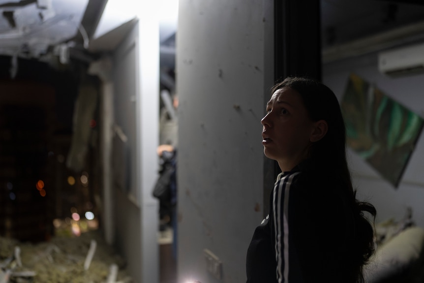 A young woman surveys her neighbour's damaged apartment in the dark. 