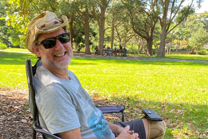 Man wearing a t shirt and shorts as well as sunglasses and hat sits in a camping chair outdoors, smiling