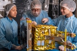 Three scientists wearing blue PPE look at a gold machine