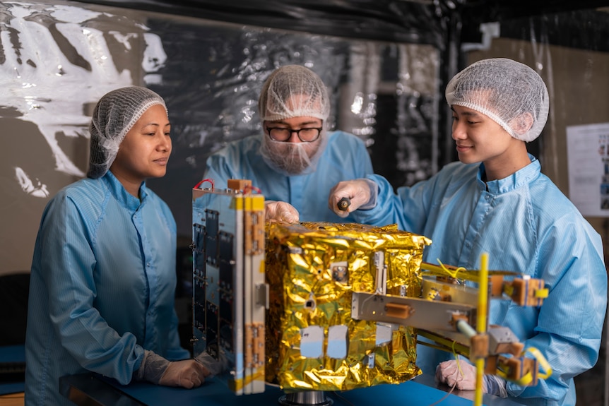 Three scientists wearing blue PPE look at a gold machine