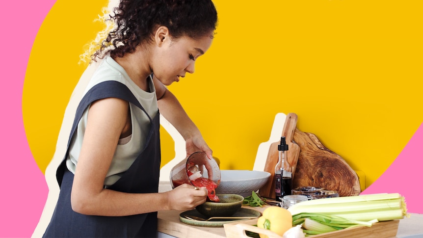 A woman wears apron and pours puree into a bowl surrounded by cutting boards, condiments and fresh produce