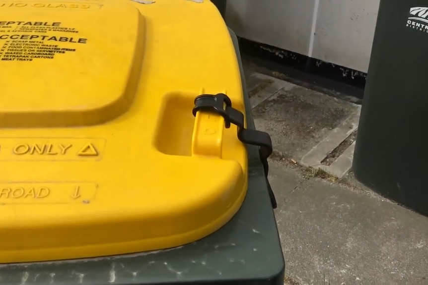 A yellow lidded bin with a black plastic strap holding on to one of the lid's handles
