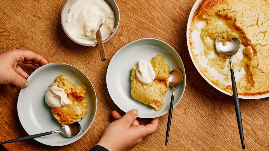 Two bowls of lemon delicious topped with cream, with the baked lemon pudding and serving cream sitting along side them.