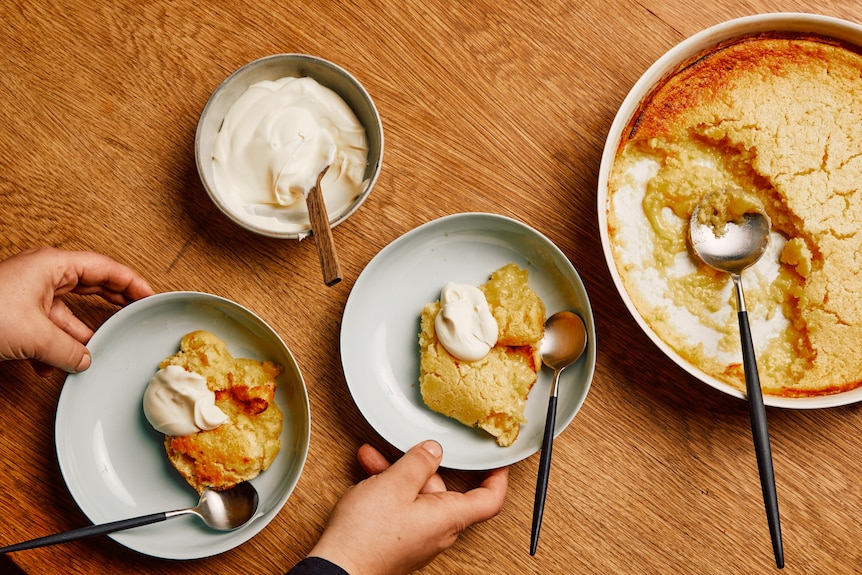 Two bowls of lemon delicious topped with cream, with the baked lemon pudding and serving cream sitting along side them.