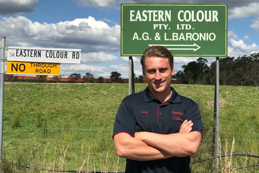 A smiling man with neatly combed dark hair stands with his arms folded in front of a paddock.