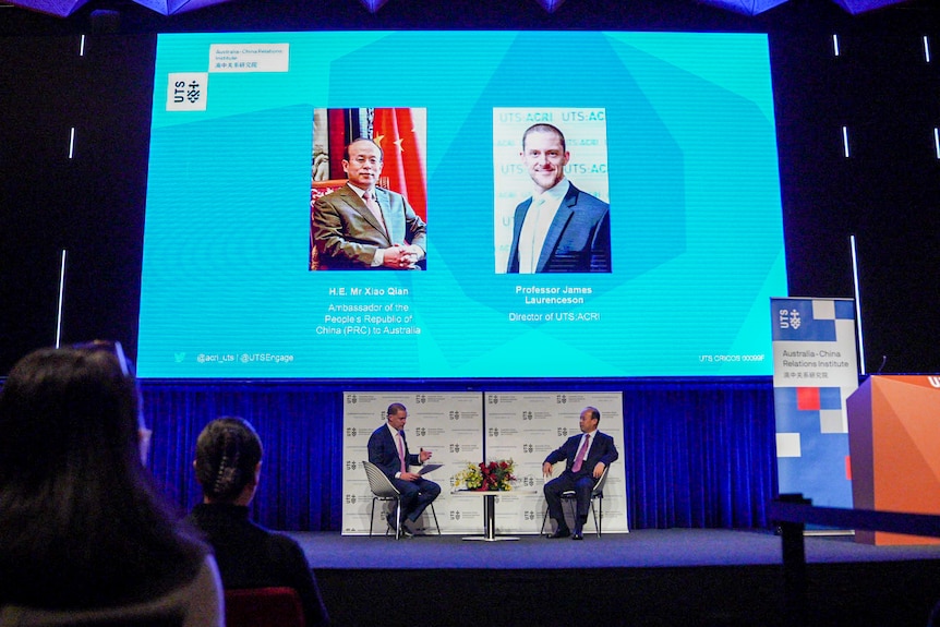 Two men sitting on a stage with their headshots projected above them on a screen.