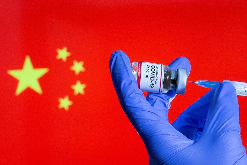 A blue glove holding a small bottle of coronavirus vaccine with a syringe with a blurry Chinese flag in the background.