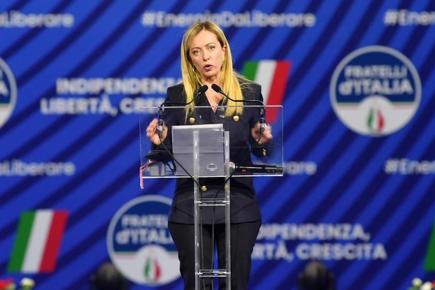 Giorgia Meloni dressed in a dark suit stands at a podium with a display of Brother of Italy behind her.