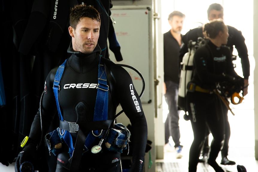 front left of frame man in scuba diving gear, wetsuit, on boat, background right other divers