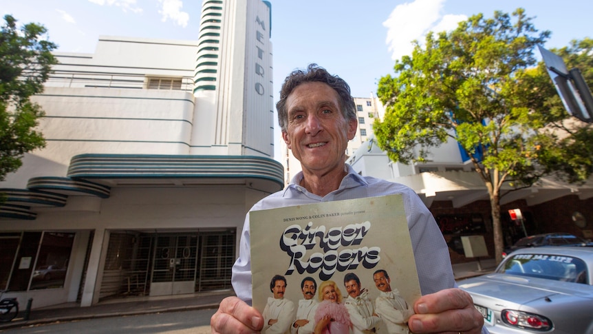 A middle-aged man holds a Ginger Rogers program, in front of an art deco theatre.