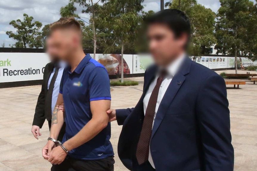 Two men wearing suits hold a man in handcuffs wearing shorts and a T-shirt