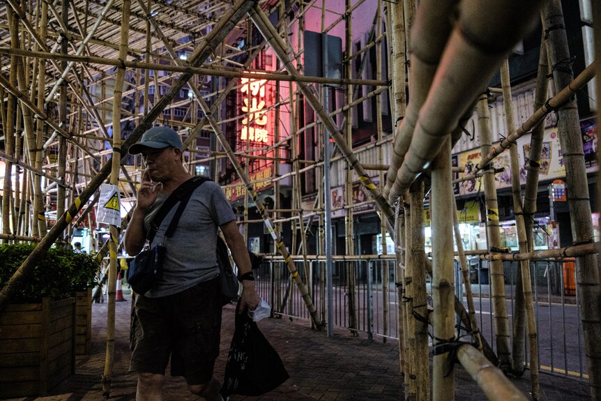 An older man in grey baseball cap smokes in front of scaffolding and Chinese signage in city