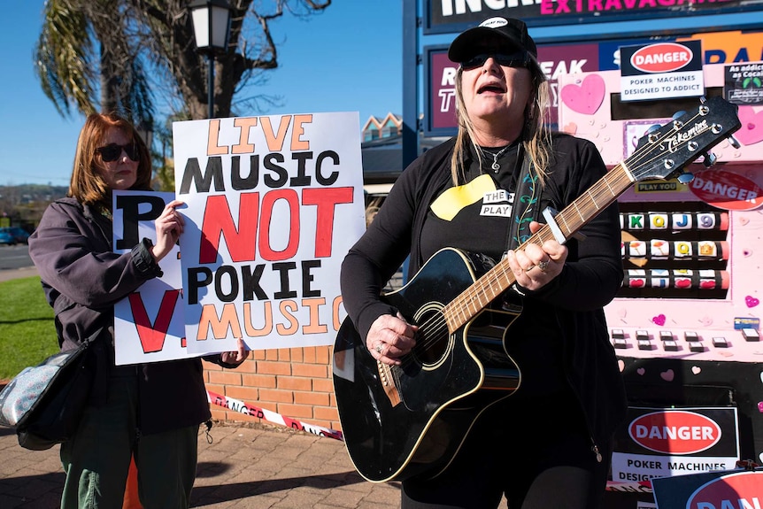 A woman plays a guitar in front of a placard saying, live music not pokie music.