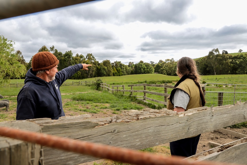 Older man wearing beanie, standing next to woman in vest, points to a green paddock from a farm lot