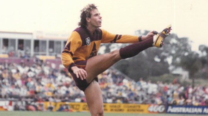 Man with blonde hair kicking in VFL Brisbane Bears uniform on a field in front of an crowd.