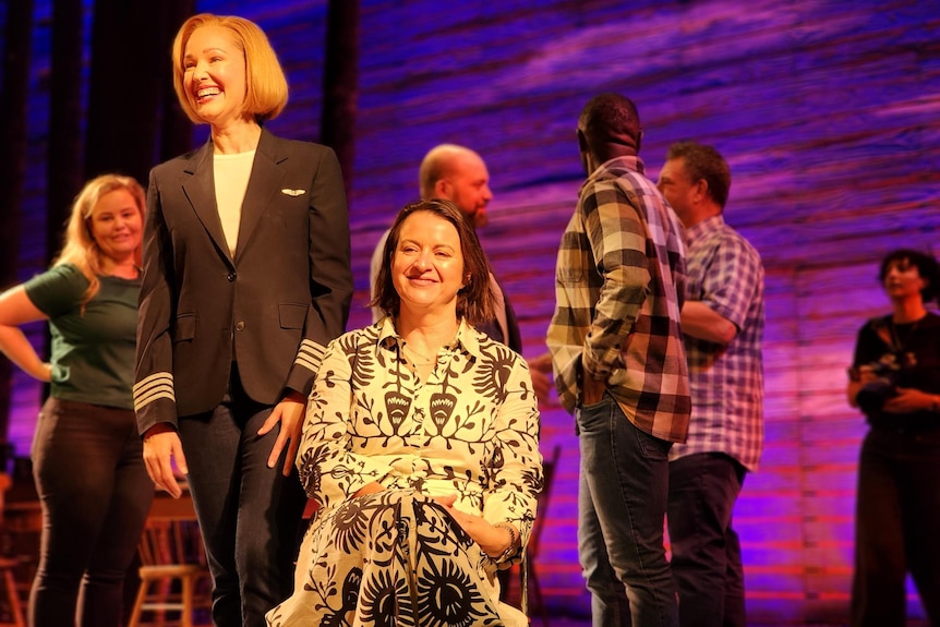 A woman seated and a woman in a pilot uniform stands next to her. Theatre cast members behind them on stage