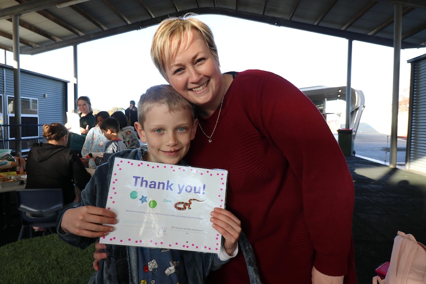 A young boy and his Mum in a red jumper hold up a thank you sign smiling.
